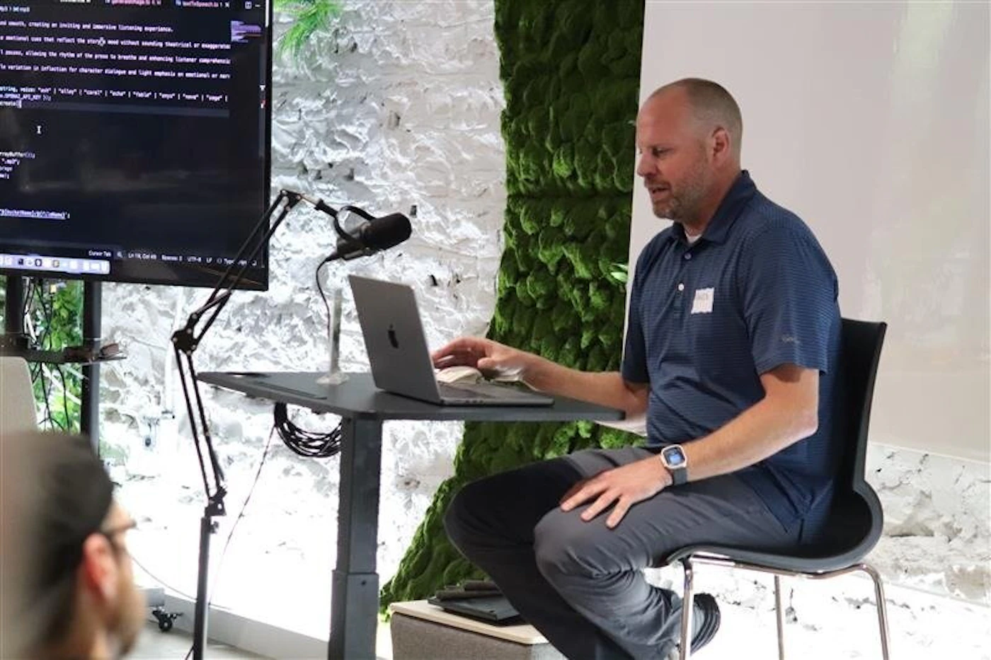 (Photo) A man in a blue polo shirt is seated at a table with a laptop, looking towards a large monitor displaying code or text. Another person's head is partially visible in the foreground, suggesting an audience or collaborative setting. The background features a distinctive textured wall with sections of green moss and white, giving the space a modern, natural aesthetic. Text: smooth, creating an inviting and immersive listening experience. elements that reflect the story's mood without sounding theatrical or exaggerated. allowing the rhythm of the prose to breathe and enhancing listener comprehension. variation in inflection for character dialogue and light emphasis on emotional or key waiting, 'water' | 'key' | 'delay' | 'reverb' | 'halo' | 'keys' | 'page' | 'wage'} PRIMARY_KEY city Cursor Talk Line 19 Col 14 UTF-8 LF TypeScript TSLint people and technology in a modern setting | indoor, likely an office, conference room, or event space | A man presenting or working with a laptop and a large screen, a microphone, a textured wall with moss, and a partially visible audience member. | candid, event or presentation photography | Colors: #335577, #666666, #99AA99, #FFFFFF Note: This image is a realistic capture of a scene, showing people, objects, and an environment as they appear in reality, which is characteristic of a photograph.