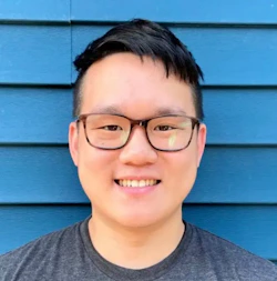 (Photo) A close-up photo of a young man wearing glasses, smiling at the camera. person | outdoor | glasses, smiling | portrait | Colors: #2f72bd, #414141 Note: The image is a real-life captured image of a person, making it a photograph. It is not a drawing, logo, or other graphic type.