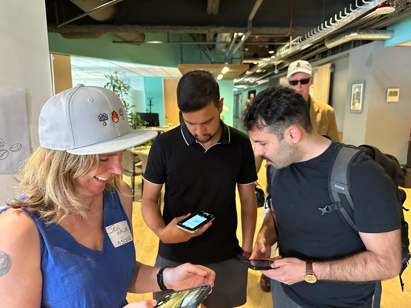 (Photo) A group of people are looking at their phones in an office setting. One person is wearing a name tag. Text: JEN HALLIE - ASCEND people | indoor | group of people, office environment | candid Note: This is a candid photograph of people in a real-world setting. It captures a moment in time and is not a logo, illustration, banner, or other graphic type.