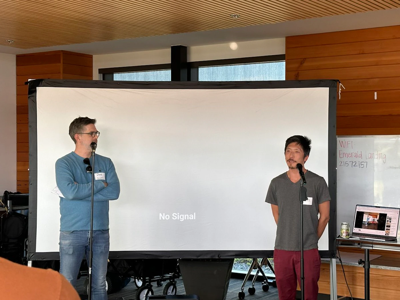 (Photo) Two men stand in front of a large projection screen. One man is wearing a blue sweater and jeans, while the other wears a gray t-shirt and red pants. They both have microphones. Text: No Signal WIFI Emerald Landing 215 72 157 people | indoor | Two men with microphones, projection screen, whiteboard | candid Note: The image is a real-life captured scene of people in a room with a projector, clearly a photograph and not a digitally created image.