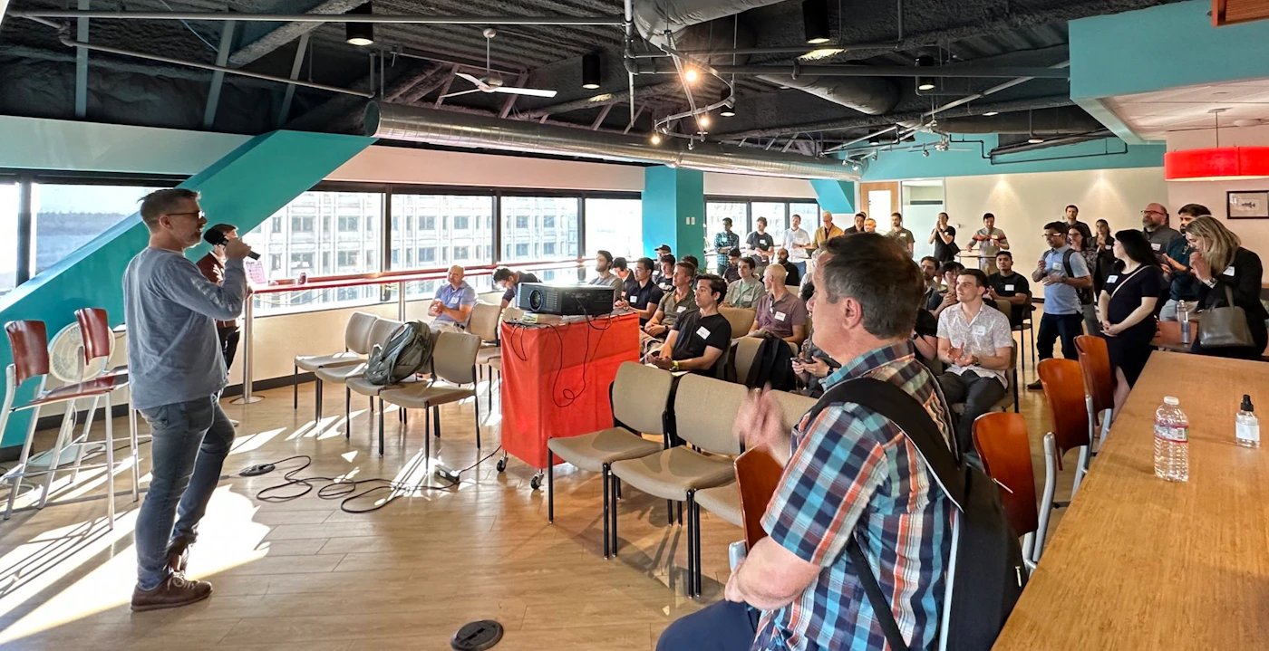 (Photo) A man speaks into a microphone to a seated audience in a large room with windows. people | indoor | presentation, audience, large room | candid Note: This is a real-life captured image of a presentation or meeting, depicting people and a scene, thus categorizing it as a photograph.