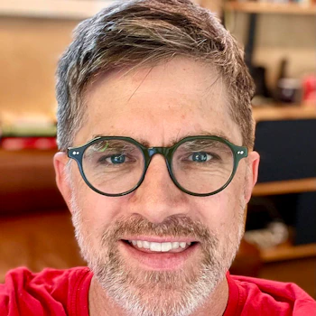 (Photo) A close-up portrait of a smiling man with short, salt-and-pepper hair, a beard, and green-framed glasses. person | indoor | green glasses, beard, red shirt, smiling | portrait | Colors: #e11d24, #2c3e2e, #d9b49e, #6b5c54 Note: The image is a realistic photograph capturing a person's face and upper torso in a portrait style.