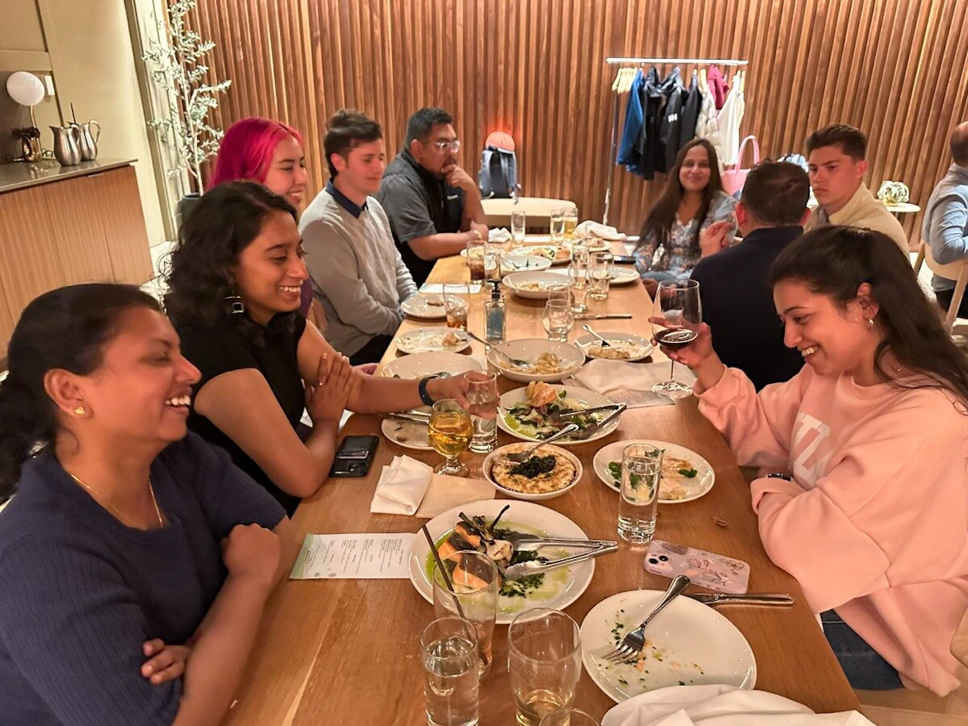 (Photo) A candid photo captures a diverse group of people, appearing to be friends or colleagues, gathered around a long wooden table in a warmly lit restaurant, enjoying a meal and conversation. A group of people dining and socializing. | Indoor, a restaurant or dining establishment. | Multiple people smiling and interacting, a long wooden table with plates of food and drinks, warm ambient lighting, a clothing rack visible in the background. | Candid, group shot, casual. | Colors: #8B5E3C, #F8C8B8, #2C3E50, #D4A78A, #F0F0F0 Note: The image is a direct capture of a real-world scene featuring multiple individuals engaged in a social activity, characteristic of a photograph.