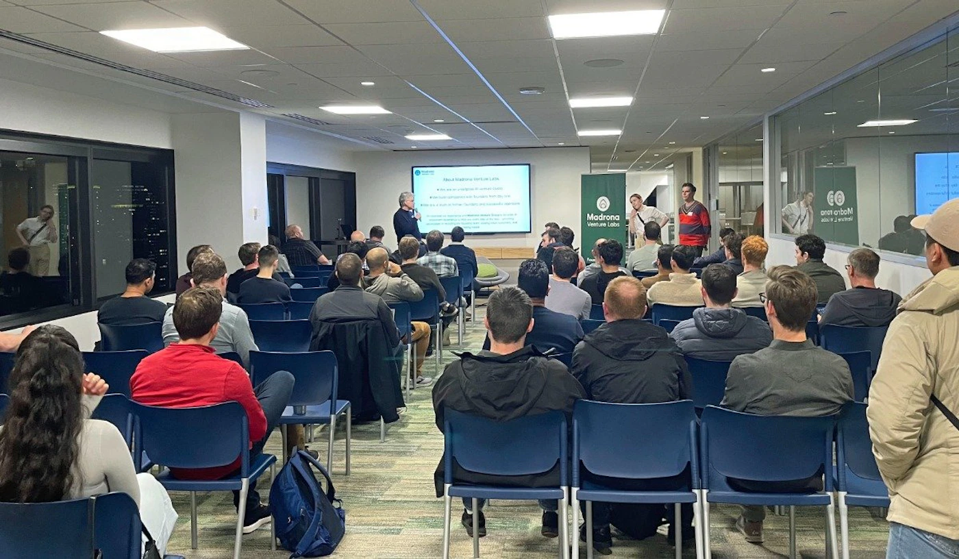 (Photo) A photo of a presentation taking place in a conference room. An audience sits in chairs facing a speaker and a projected presentation. people | indoor | presentation, audience, speaker | candid Note: The image is a real-life captured image of a scene, showing a presentation in progress. This makes it a photograph.