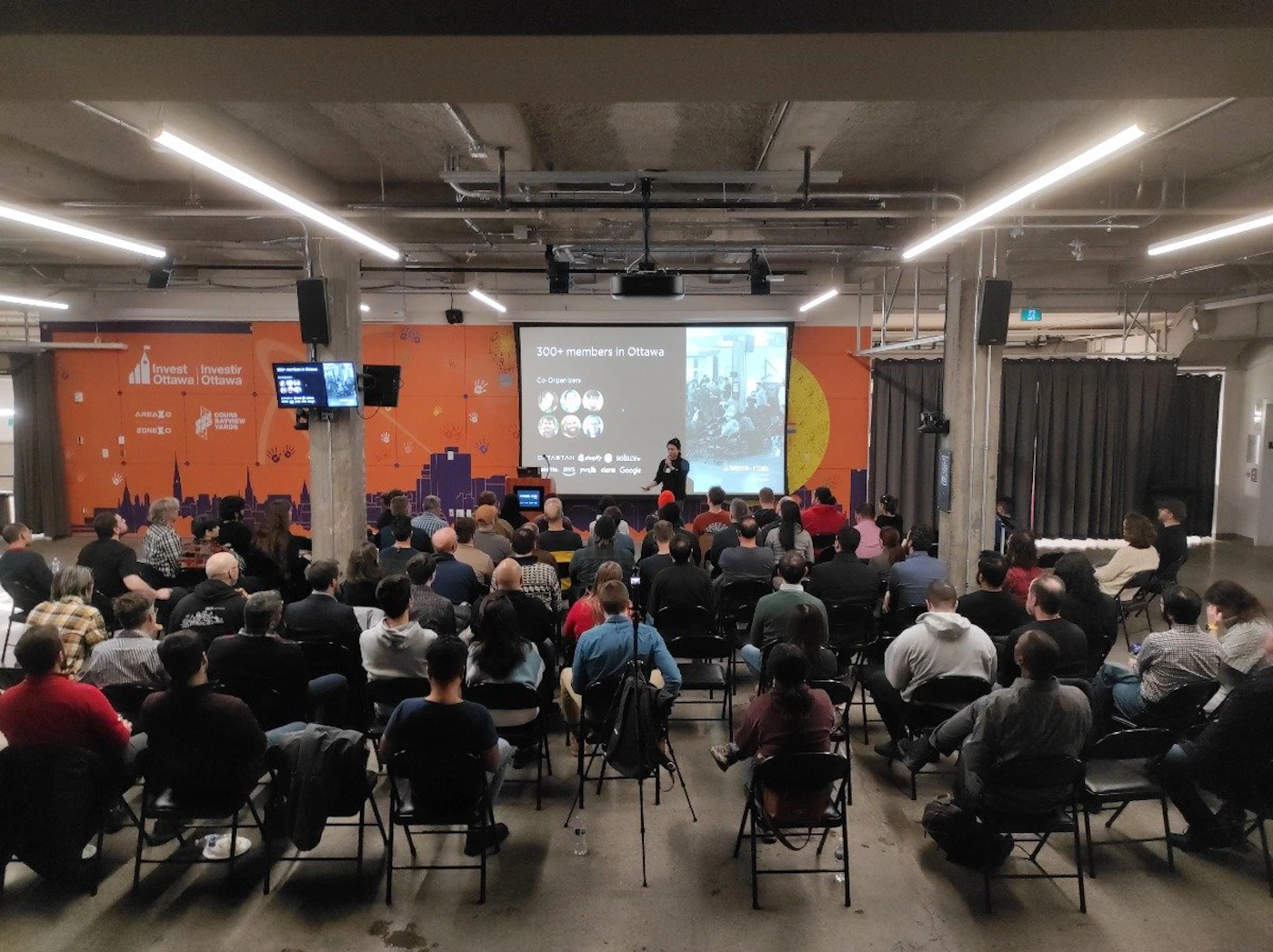 (Photo) A photo of a presentation taking place in a large room. An audience sits in folding chairs facing a projector screen, with a speaker presenting near the screen. Text: 300+ members in Ottawa Go deeper into: [unreadable] people | indoor | presentation, audience, projector screen | candid Note: The image is a real-life capture of an event, depicting a presentation with an audience. This clearly falls under the category of a photograph.