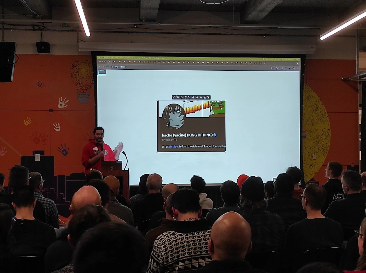 (Photo) A man in a red hoodie is giving a presentation to a seated audience. He stands at a podium in front of a large screen displaying a social media post. Text: kache (yacine) (KING OF DING) @yadine/TB Al, ex stripe, follow to watch a self funded founder be ACL person | indoor | presentation, audience, projector screen | candid Note: The image is a real-life captured moment of a presentation, making it a photograph. It's not a staged image or other visual media type.