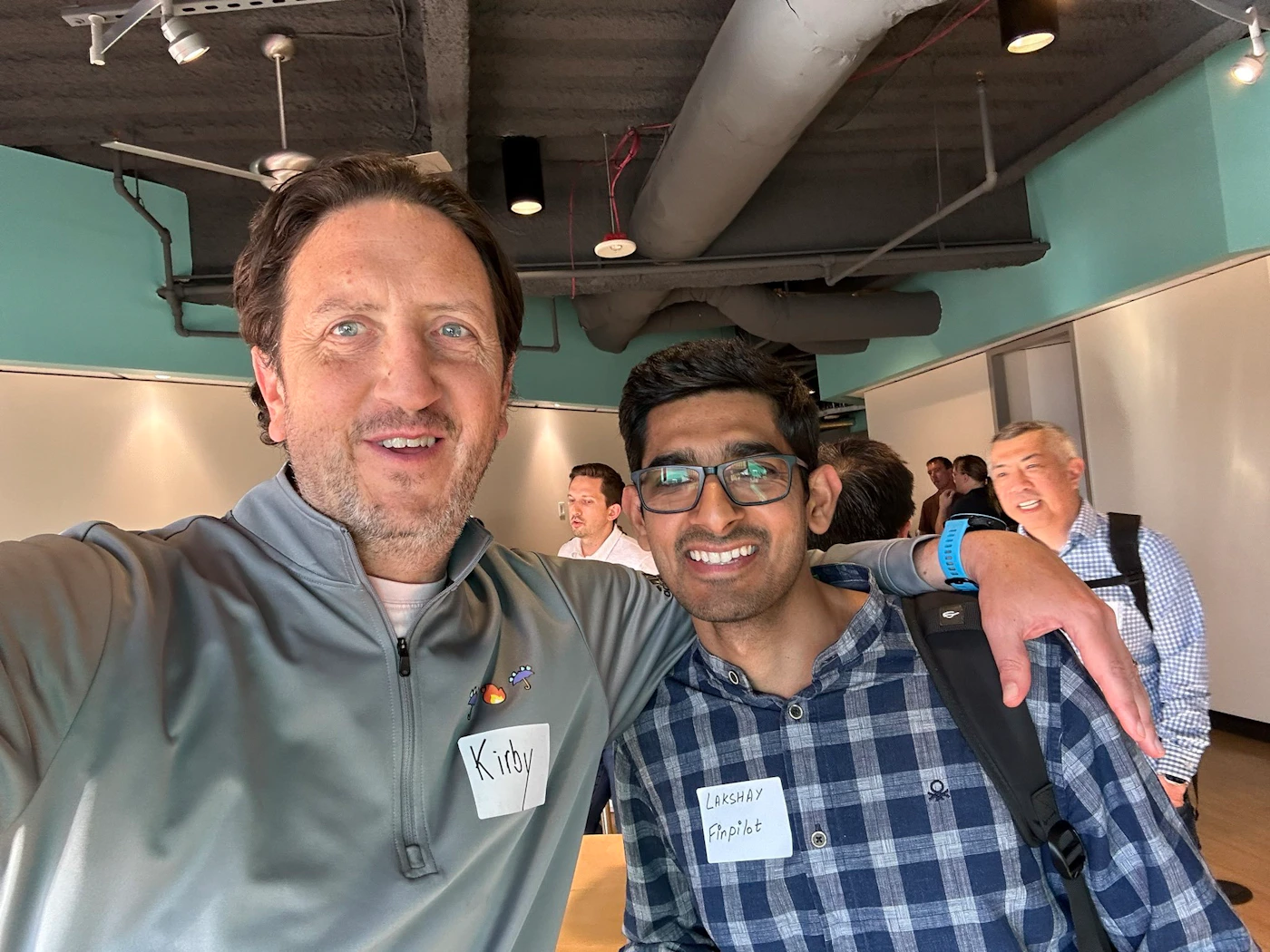 (Photo) A selfie of two men, one older and one younger, wearing name tags at what appears to be a casual work event. Text: Kirby Lakshay FinPilot person | indoor | multiple people, name tags, casual attire | selfie Note: The image is a real-life captured image of people in an indoor setting, indicative of a photograph. It is not a drawing, logo, or banner.
