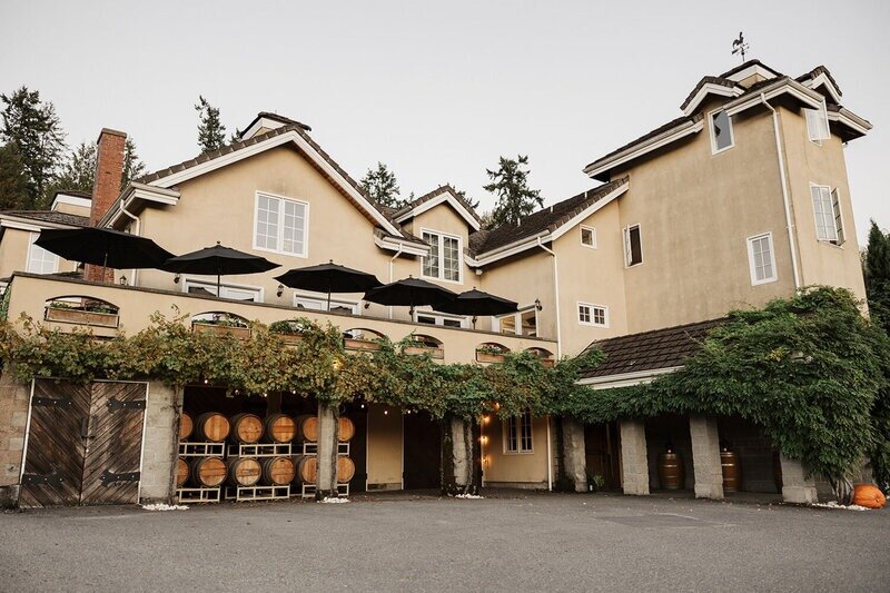 (Photo) A photograph of a large, multi-story building, likely a winery or event venue, featuring tan stucco walls and dark tiled roofs. Architectural structure (winery/venue) and wine barrels | Outdoor, likely a vineyard or forested area, under an overcast sky | Lower level storage area displaying stacked wooden wine barrels, a terrace with black patio umbrellas, extensive green vines covering the facade, and a weather vane on the roof. | Environmental/architectural photography, slightly muted colors | Colors: #CFC0A7, #55453E, #3E543C, #999999 Note: The image is a realistic capture of an architectural structure (a building) and its environment, classifying it as a photograph.