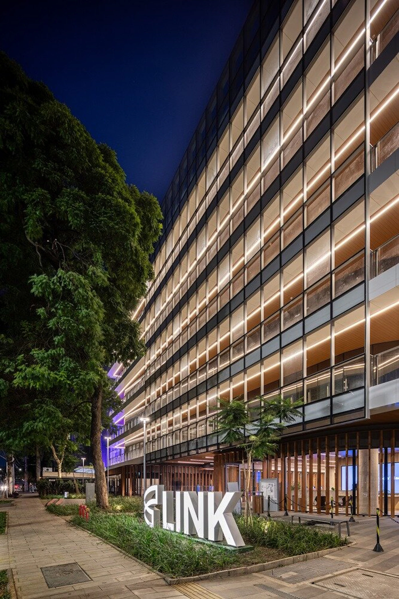 (Photo) A nighttime photograph showcasing a modern multi-story building with a glass facade and warm interior lighting, featuring a large illuminated 'LINK' sign prominently displayed in the foreground amidst green landscaping. A sidewalk and trees are visible on the left, under a dark blue night sky. Text: LINK Modern building, urban environment, architectural structure | Outdoor, urban, night | Large illuminated 'LINK' sign, multi-story glass facade with horizontal strip lighting, lush green trees, paved sidewalk, dark night sky. | Architectural photography, night scene | Colors: #0A1128, #2C5900, #F5E8C7, #A0A0A0, #FFFFFF Note: The image is a realistic capture of a real-world scene, specifically an architectural view of a building and its urban surroundings at night, which falls under the 'photo' category.