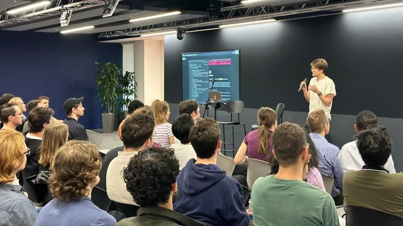 (Photo) A person stands at the front of a room, speaking into a microphone to a seated audience.  A screen behind the speaker displays lines of code. people | indoor | presentation, audience, screen with code | candid Note: This is a real-life captured image of a presentation, depicting people and a setting, making it a photograph.