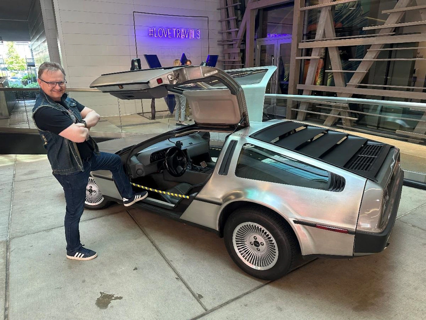 (Photo) A man poses with one foot on a DeLorean car with gull-wing doors open. Text: #LOVETRAVELS person, car | indoor | DeLorean car, gull-wing doors open | candid Note: The image is a real-life photograph of a person and a car. It captures a moment in time and is not a drawing, logo, or banner.