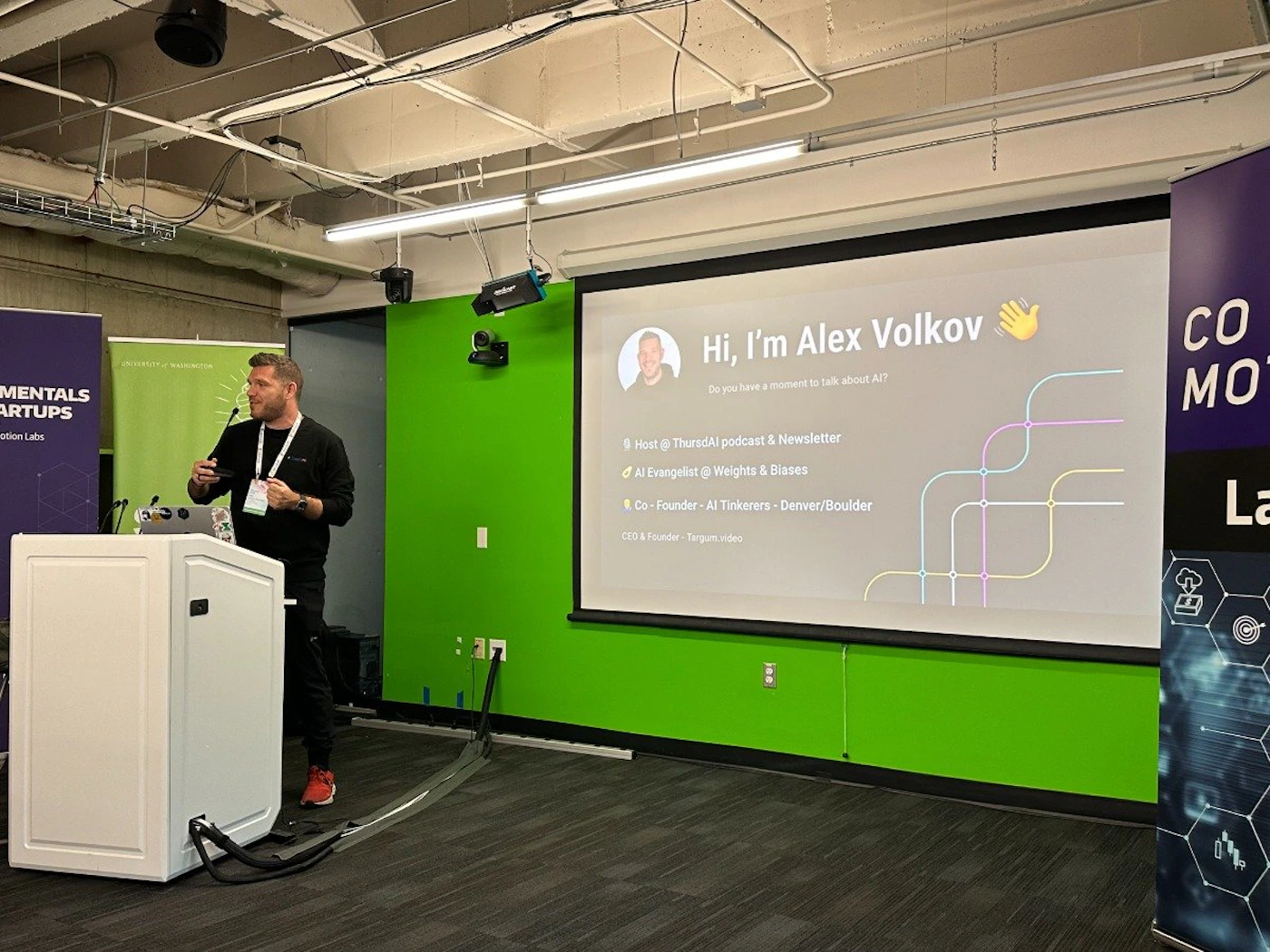 (Photo) A man stands at a podium giving a presentation about AI in a room with a green wall and a projected slide. Text: Hi, I'm Alex Volkov Do you have a moment to talk about AI? Host @ ThursdAl podcast & Newsletter AI Evangelist @ Weights & Biases Co-Founder - AI Tinkerers - Denver/Boulder CEO & Founder - Targum video MENTALS ARTUPS Motion Labs CO MO La person | indoor | presentation, projector screen, podium | candid Note: The image is a real-life captured image of a presentation, making it a photograph. It's not a logo, illustration, or other specific types.