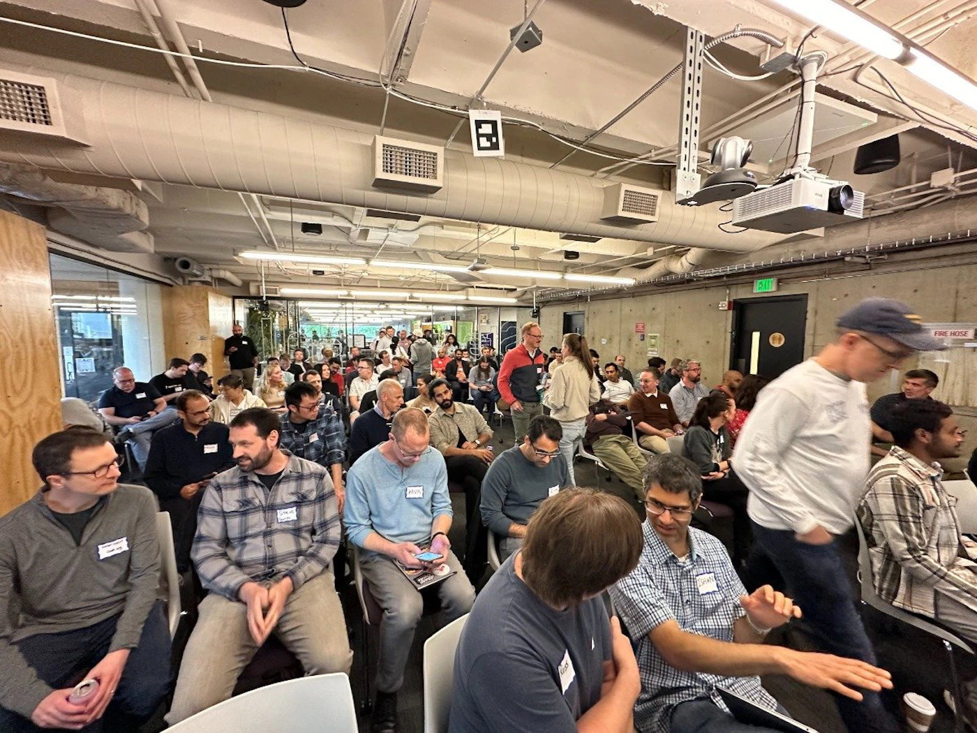 (Photo) A photo of a large group of people sitting and standing in a room, seemingly attending a presentation or meeting. people | indoor | large group, presentation setting | candid Note: The image is a real-life captured image of a gathering of people in an indoor setting.  It exhibits characteristics of a candid photograph.