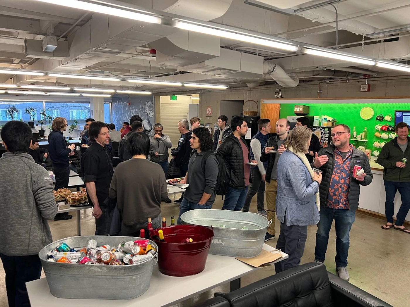 (Photo) A photo of a group of people gathered in an office space, possibly for a social event or casual gathering. people | indoor | group of people, office setting, food and drinks | candid Note: The image is a real-life captured scene of people in an indoor environment, clearly a photograph rather than a drawing or digitally created image.  There are no textual elements designed as part of the image itself.