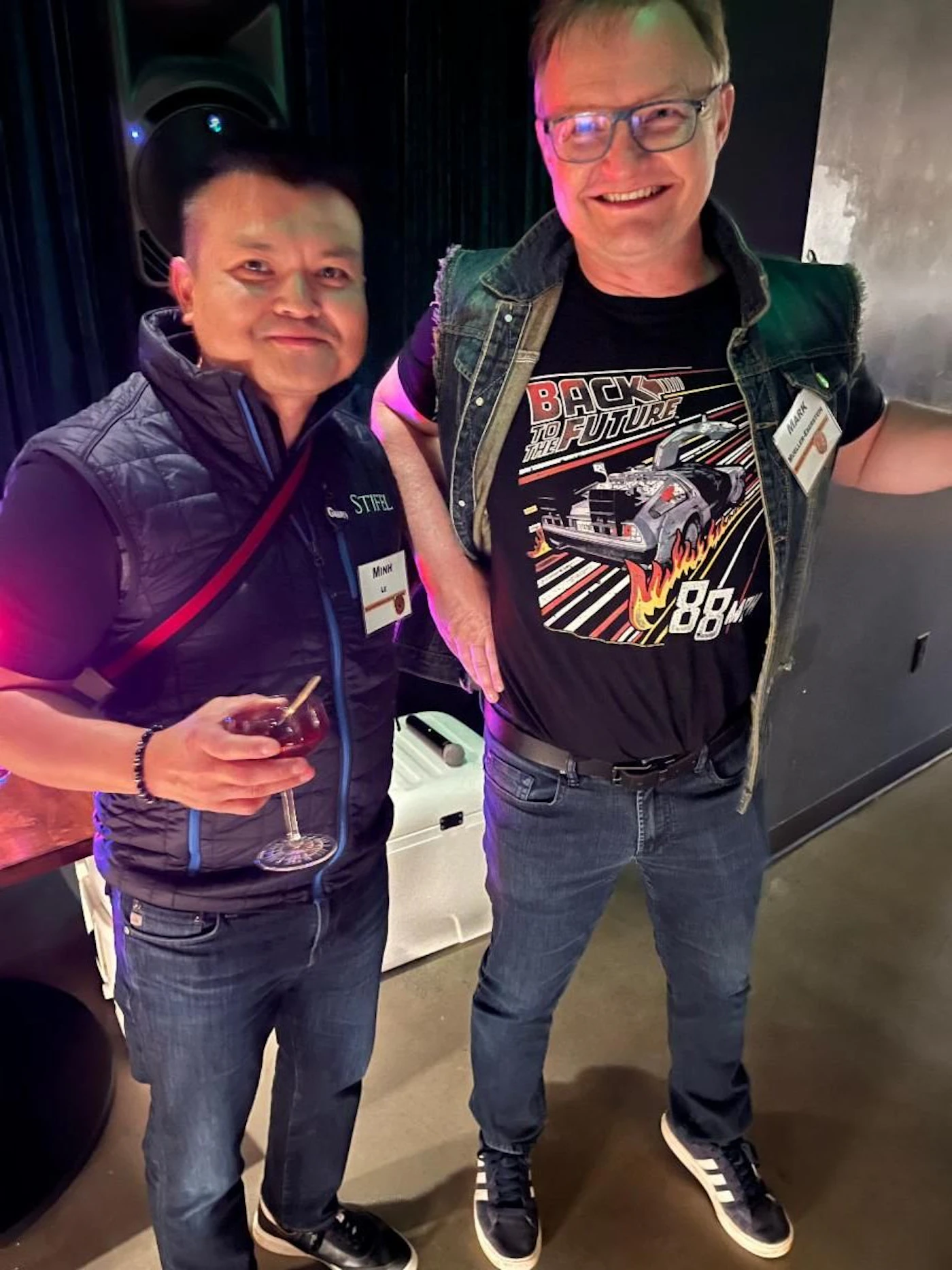 (Photo) Two men pose for a photo, one holding a drink. The man on the right wears a Back to the Future t-shirt. Text: BACK TO THE FUTURE 88 person | indoor | two people | candid Note: The image is a real-life captured image of two people, clearly a photograph and not a digitally created image.