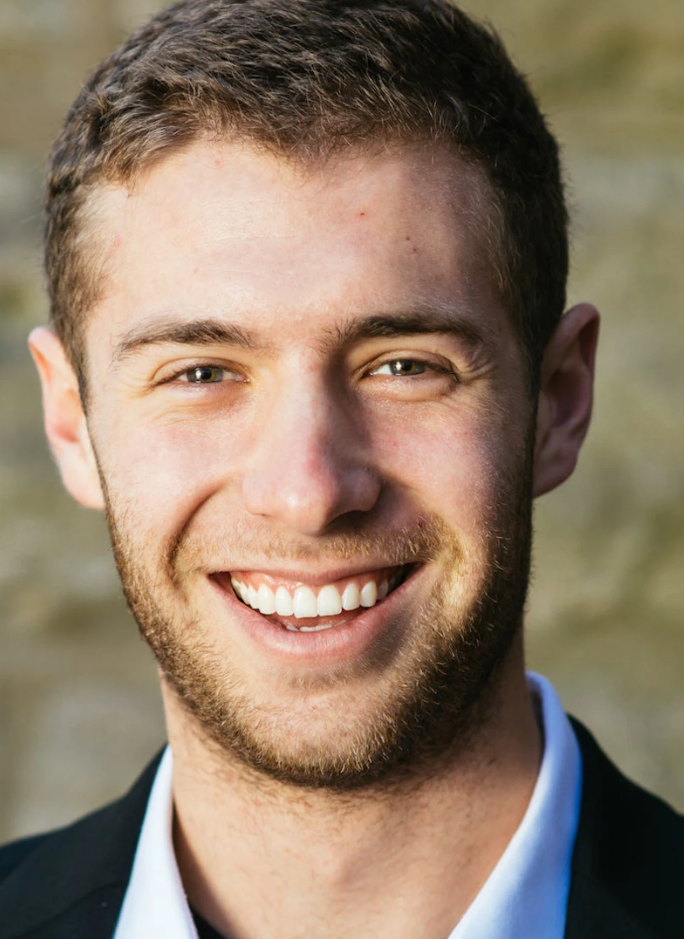 (Photo) A close-up portrait features a smiling young man with short brown hair and a beard, wearing a dark jacket over a white collared shirt. The background is softly blurred in warm, earthy tones. person | outdoor | smiling, close-up, beard, natural light | portrait, candid | Colors: #E0B494, #5C3D2B, #FFFFFF, #1A1A1A, #A3967F Note: The image is a realistic capture of a person, exhibiting the characteristics of a photograph rather than a stylized illustration, logo, banner, or handwriting.