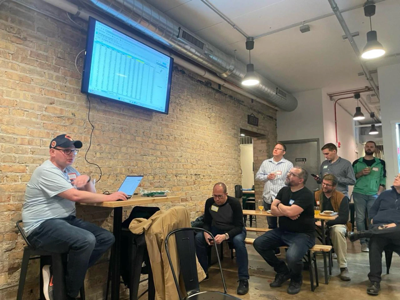 (Photo) A group of men are gathered indoors in what appears to be a casual meeting or presentation setting. One man is seated at a high table with a laptop, facing a large screen displaying a spreadsheet, while other attendees stand or sit, observing the screen or the presenter. A group of people attending a presentation or meeting | Indoor, in a venue with exposed brick walls and an industrial aesthetic, possibly a co-working space or event room. | A large wall-mounted monitor displaying unreadable spreadsheet data, a man presenting from a laptop, several men observing, and exposed ceiling pipes and industrial lighting. | Candid, event photography | Colors: #8B6D5A, #E0E0E0, #333333, #90C0D0 Note: The image is a direct capture of a real-world scene featuring people and an indoor environment, fitting the definition of a photograph.
