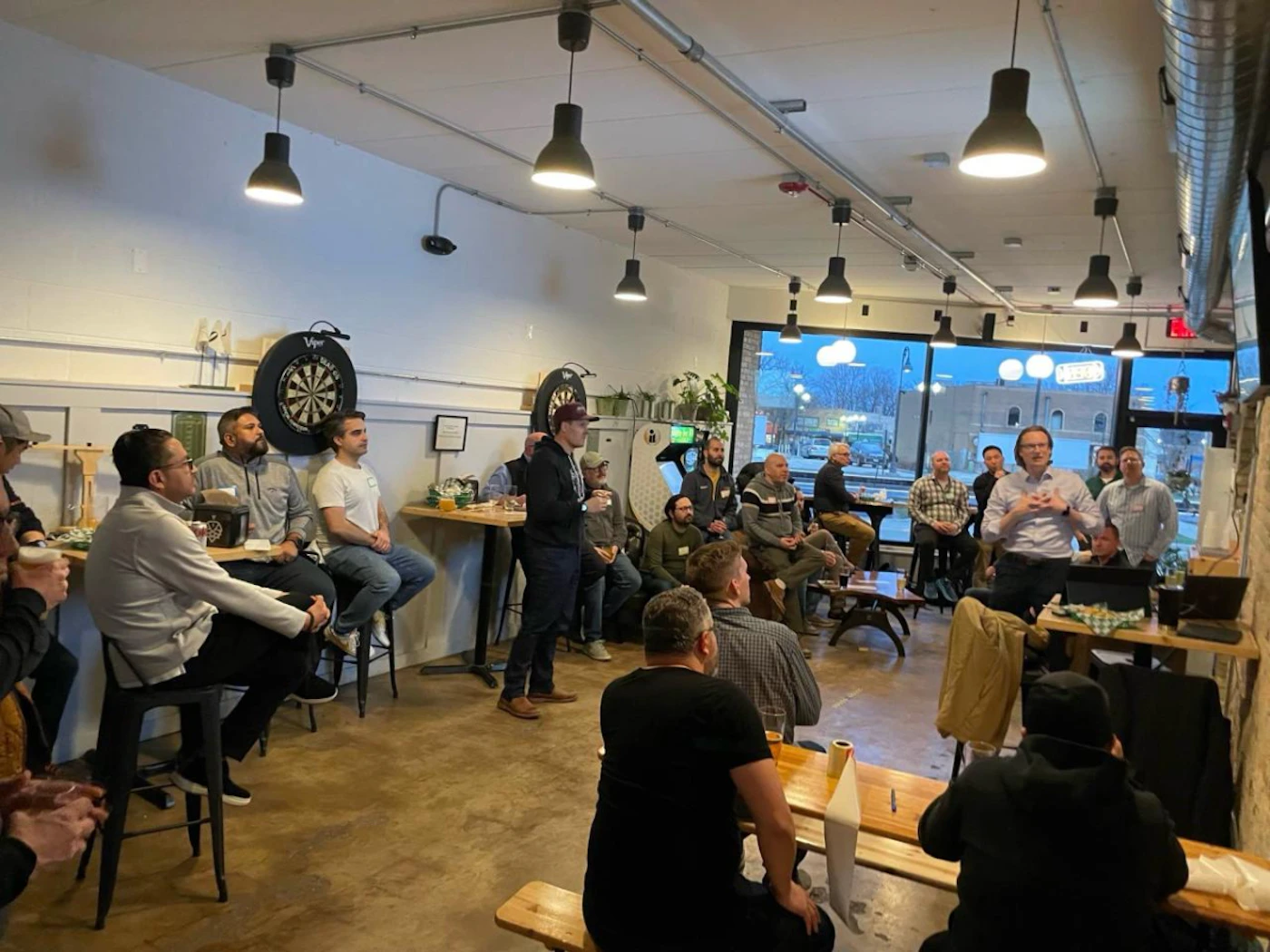 (Photo) A group of men are gathered indoors in what appears to be a casual venue, possibly a bar or a community space. One man is standing in the center, speaking to the seated and standing attendees, under industrial-style pendant lights. A group of men attending a gathering or presentation | Indoor, casual venue (possibly a bar or event space) with large windows showing an outdoor street scene at dusk/night. | Multiple people (mostly men), one person speaking, dartboards on the wall, high tables and stools, industrial pendant lighting, exposed ductwork. | Candid, indoor event photography. | Colors: #E0E0E0, #705541, #3A3A3A, #A0B0C0, #FFFFE0 Note: This image is a direct capture of a real-world scene involving people and an indoor environment. It depicts a moment in time rather than being a graphic design, logo, or illustration.