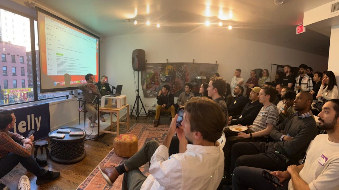 (Photo) A photo of a presentation or talk in a room with an audience.  A presenter is on a small stage with a projected screen behind them, and the audience is seated in chairs. Text: Jelly people | indoor | presentation, audience, projector screen | candid Note: The image is a real-life captured scene of an event with people, making it a photograph. It is not a drawn or digitally created image.