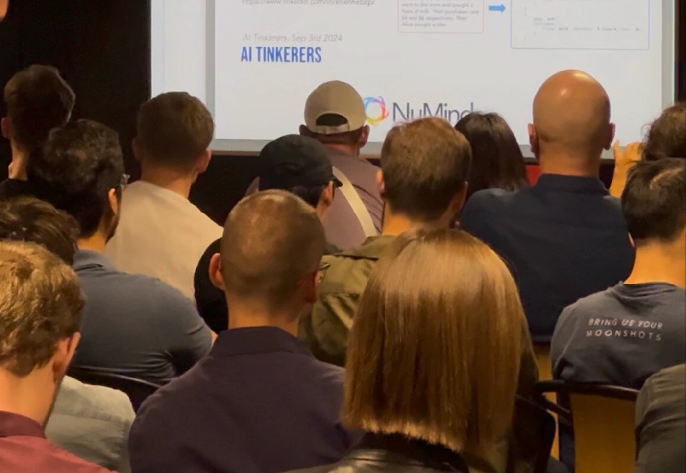 (Photo) The image captures a view from behind an audience attending a presentation, with a projector screen visible in the background displaying text and a logo. Many individuals are seated, looking towards the screen, suggesting a conference or lecture setting. Text: https://www.linkedin.com/in/etiennebcp/ went to the store and bought 1 type of milk. Their purchases cost $1 and $2 respectively. Then Alice bought a bike. AI Tinkerers, Sep 3rd 2024 AI TINKERERS NuMind BRING US YOUR MOONSHOTS audience, presentation, people, event | indoor, likely a conference room or auditorium | multiple people viewed from behind, projector screen with text and a logo, text on a person's shirt | candid, documentary | Colors: #2C354E, #6B8BA6, #A07452, #F0F0F0, #5C4336 Note: The image is a realistic capture of a moment or scene, depicting people and an environment as they appear in real life, which is characteristic of a photograph.