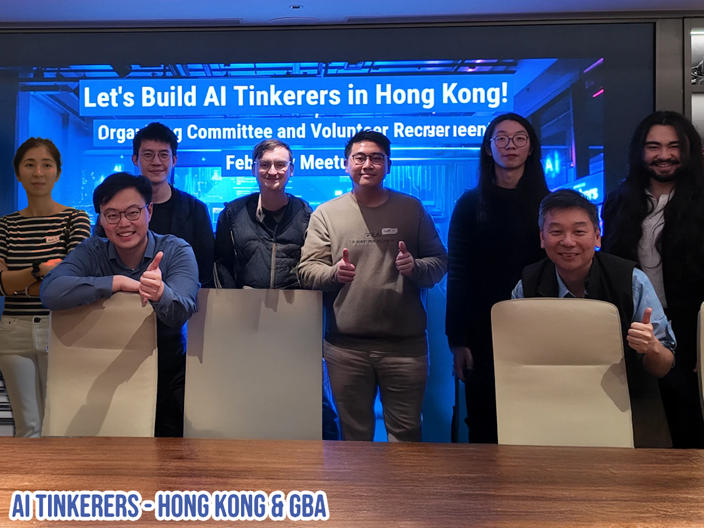 (Photo) A group of people posing for a photo in front of a screen displaying "Let's Build AI Tinkerers in Hong Kong!". The group appears to be associated with the AI Tinkerers organization. Text: Let's Build Al Tinkerers in Hong Kong! Orga Committee and Volunteer Recrser leen Feb Meetu AI TINKERERS HONG KONG & GBA group of people | indoor | group of people, screen with text | group portrait | Colors: #2E5984, #A0785A, #FFFFFF Note: The image is a photograph of a group of people. It captures a real-life scene with identifiable subjects.