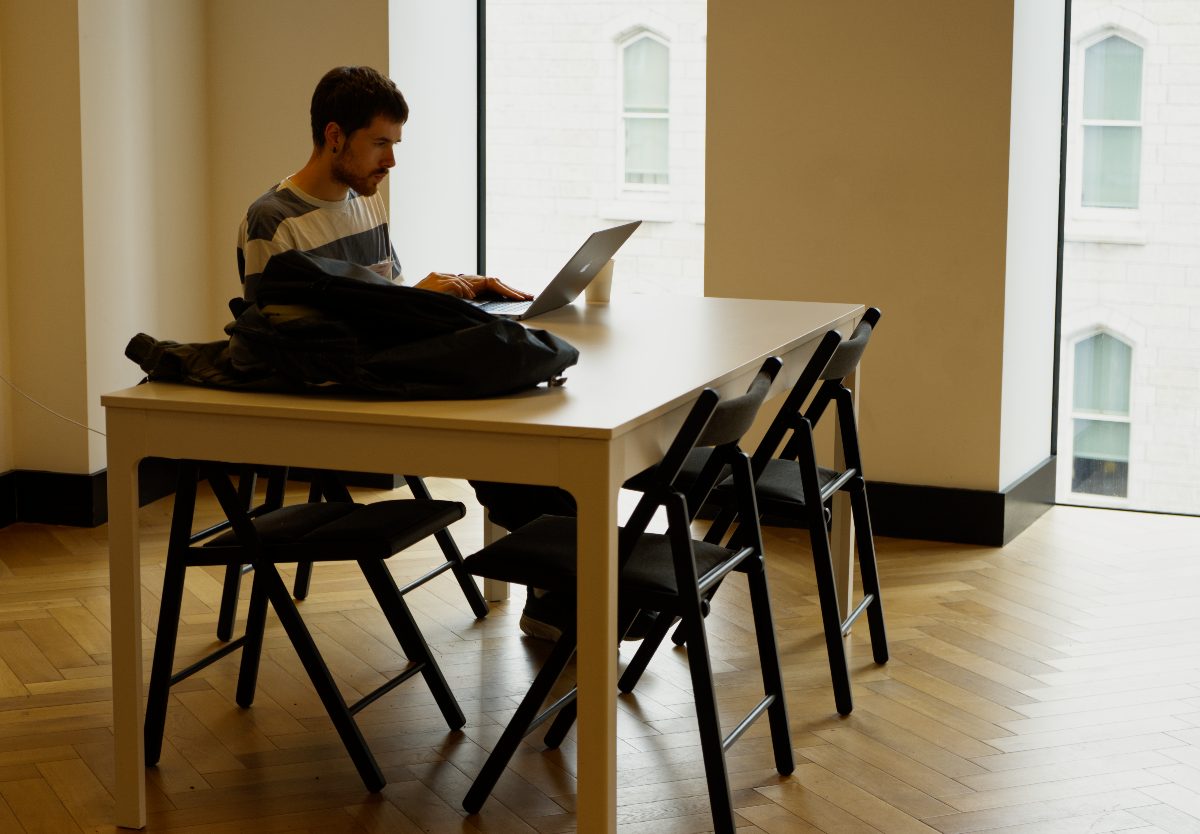 (Photo) A man with a beard is seated at a large white table, actively working on a laptop. Several black folding chairs are arranged around the table in a bright, modern room with large windows revealing an exterior building. person, indoor scene | indoor, modern office or co-working space | a man working on a laptop, large windows, herringbone wood floor, minimalist furniture, black jacket on table | candid, natural light | Colors: #F8F8F8, #2C2C2C, #C2A582, #D3D3D3 Note: The image is a realistic capture of a real-world scene, featuring a person and an indoor environment with natural lighting, characteristic of a photograph.