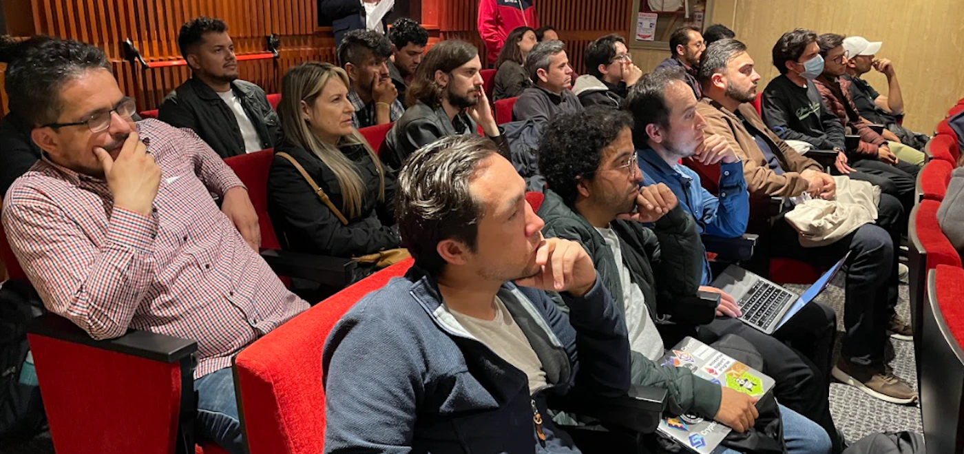 (Photo) The image shows a group of people sitting in an auditorium-like setting, possibly attending a lecture or presentation. They are seated in rows of red chairs. group of people | indoor | rows of seats, some people looking at laptops | candid | Colors: #8B0000, #D3D3D3, #000000 Note: The image appears to be a real-life photograph of a group of people, not a drawing, logo, banner, or handwriting sample.