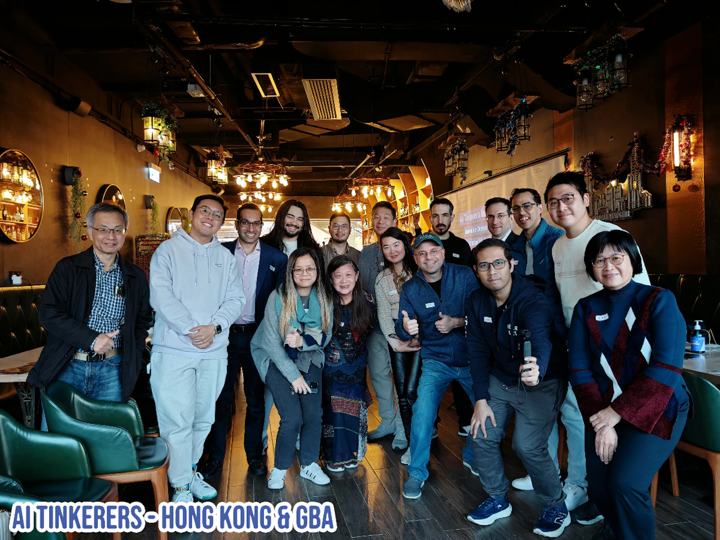 (Photo) A group of people are posing for a photo indoors. The setting appears to be a restaurant or bar. Text: AI TINKERERS - HONG KONG & GBA group of people | indoor | multiple people, restaurant/bar setting | candid | Colors: #282017, #8C725D, #FFFFFF, #003366 Note: The image shows a real-world scene with people, suggesting it is a photograph. The presence of text is secondary to the overall photographic nature of the image.
