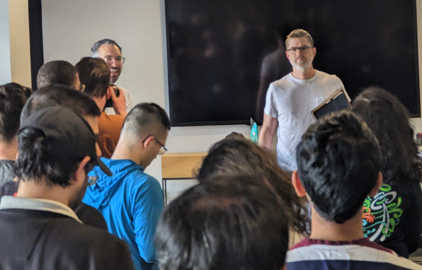 (Photo) A photo of a group of people gathered, seemingly listening to a presentation or talk. One person is standing in front of a large screen, possibly presenting, while others are facing towards him. people | indoor | group of people, presentation setting | candid Note: The image is a real-life captured image of people in a room, with no text or artistic elements suggesting other categories. Therefore, it's classified as a photo.