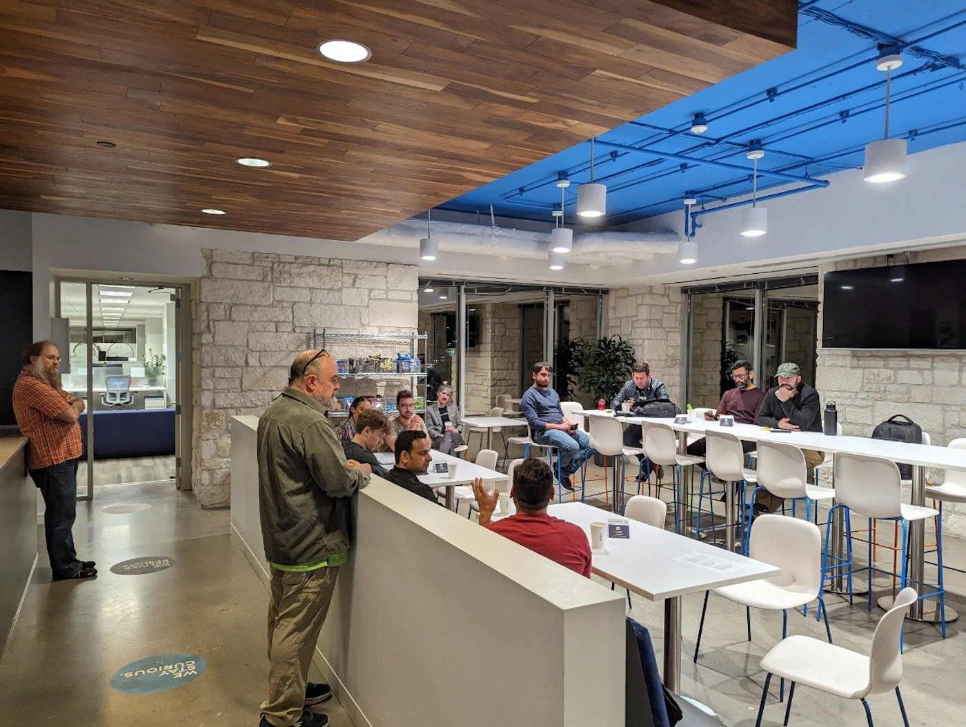 (Photo) A photo of a group of people in a modern office break room or cafeteria area.  Some are seated at tables, while others stand and talk. people | indoor | group of people, office break room | candid Note: The image is a realistic depiction of a real-world scene captured by a camera, making it a photograph.