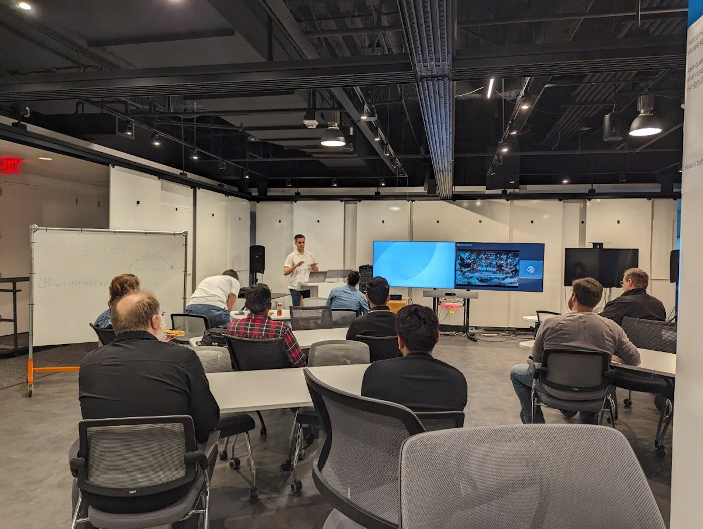 (Photo) A photo of a group of people in a presentation or training session.  A presenter stands at the front of the room, while others are seated at tables, watching a screen. people | indoor | presentation, group of people, screen | candid Note: This is a real-life captured image of a scene, depicting a group of people in a room with presentation equipment. Therefore, it's classified as a photo.
