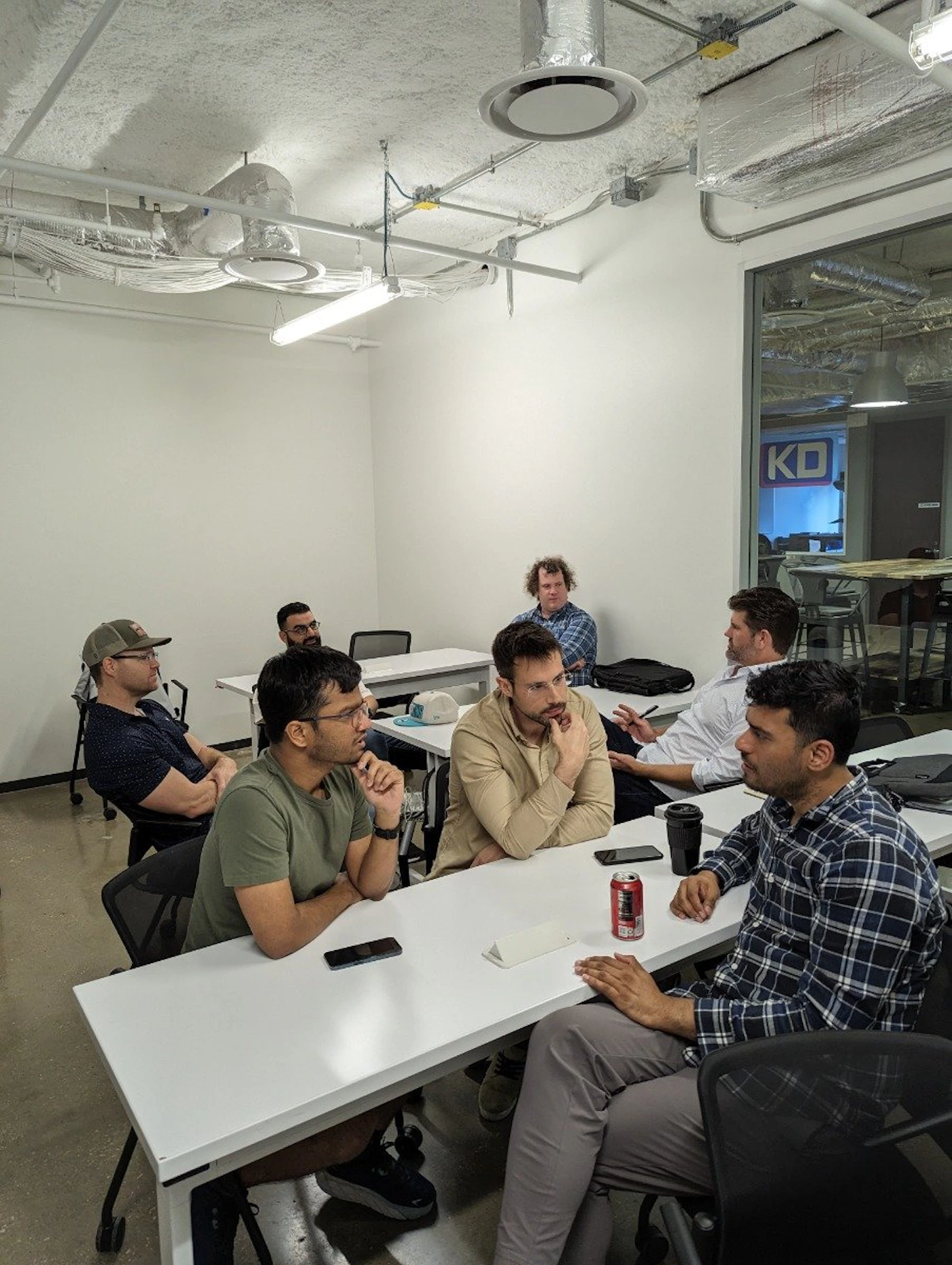(Photo) A group of men are sitting around a white table in a meeting room, seemingly engaged in a discussion. Text: KD people | indoor | group discussion, meeting room | candid Note: The image is a realistic depiction of a real-life scene captured with a camera, thus categorizing it as a photograph.