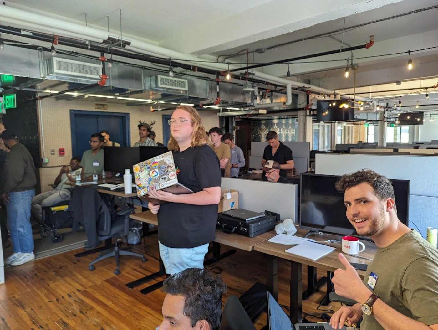 (Photo) A photo of people in an office setting, some standing and some sitting at desks. One person is holding a laptop covered in stickers. people | indoor | group of people, office environment | candid Note: The image is a real-life captured image of a scene, depicting people in a physical space, indicating it's a photograph.