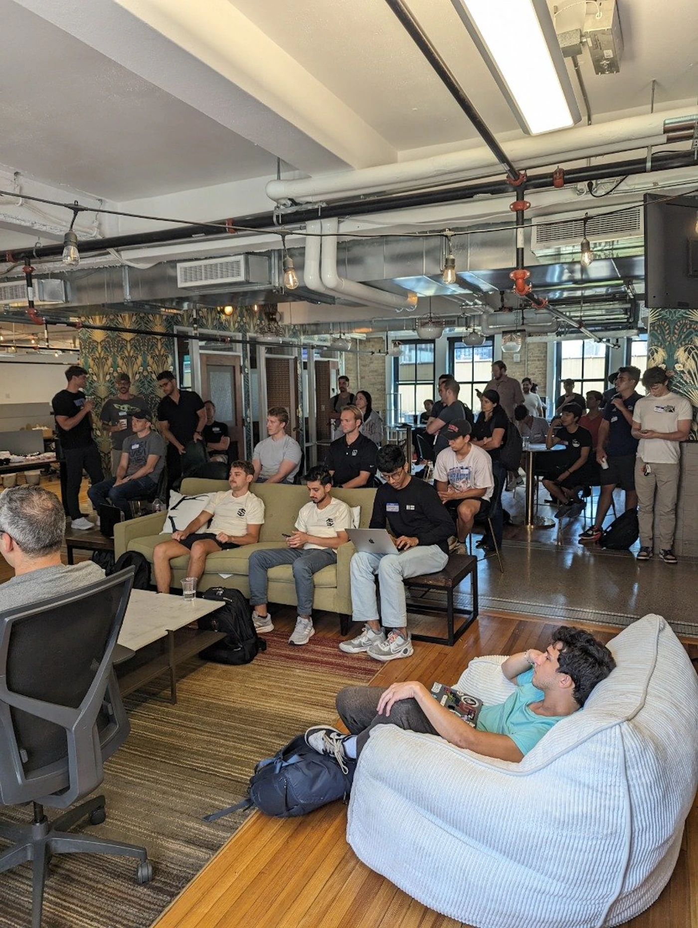 (Photo) A photo of a group of people gathered in a room, some seated on a couch and others standing, appearing to listen to a speaker. people | indoor | group of people, presentation setting | candid Note: The image is a real-life captured scene of people in a room, indicative of a photograph. It doesn't contain any drawn or digitally created elements, nor is it a logo or banner.