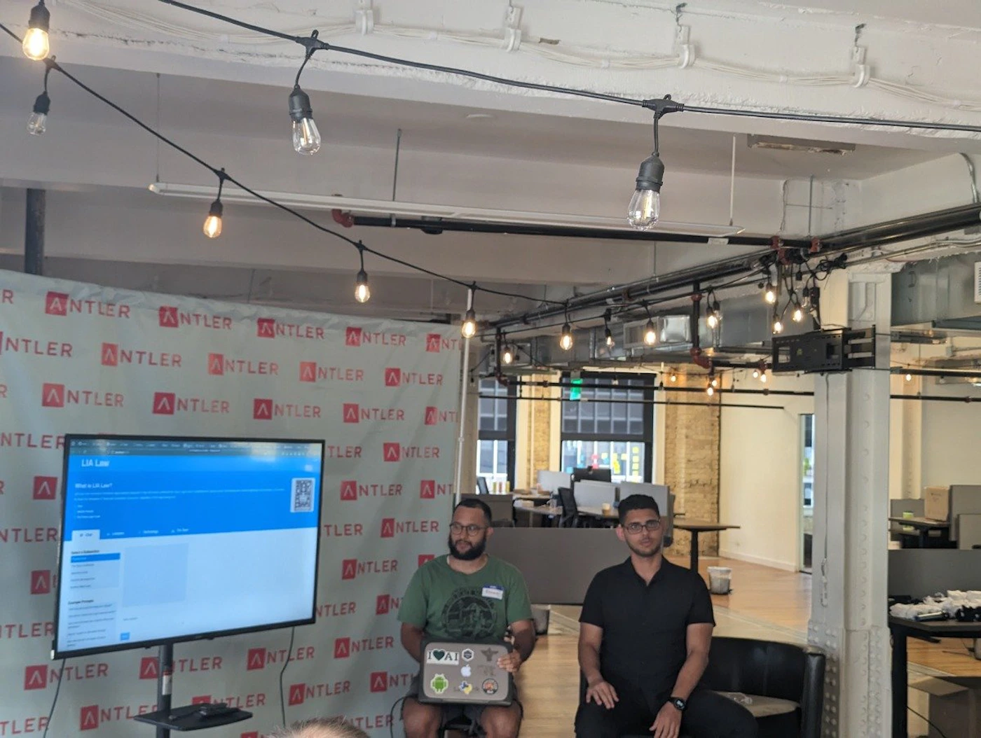 (Photo) Two men sit in front of a large screen displaying a website in what appears to be a presentation or meeting setting. The backdrop features a repeating Antler logo. Text: R ANTLER ANTLER ANTLER ANTLER INT NTLER ER ANTLER ANTLER ANTLER ANTLER ANTLER ANTLER ANTLER ANTLER ANTI R NTLER LIA Law A NTLER A TLER ANTLER TLEF TLER ANTLER ANTLER ANT TLER ANTLER ANTLER TLER AN ATLER ANTLER ANTLER ANTLER TLER ANTLER ANT TLER AL people | indoor | presentation, audience, branded backdrop | candid Note: The image is a real-life captured scene of people in an office-like environment, making it a photograph.  It includes people, a presentation screen, and branded backdrop.