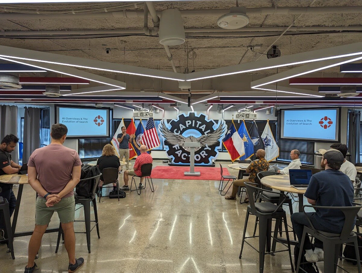 (Photo) A photo of a presentation taking place in a modern office space.  A speaker stands in front of a branded backdrop while the audience sits at tables. Text: Al Overviews & The Evolution of Search CAPITAL FATORY Al Overviews & The Evolution of Search people | indoor | presentation, audience, office space | candid Note: The image is a real-life captured scene of an indoor event, making it a photograph. It's not a logo, illustration, or other specific type.
