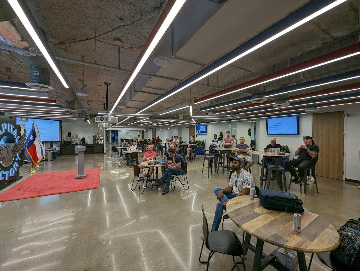 (Photo) A photo of a large room with people sitting at tables, apparently listening to a presentation.  A podium and screen are visible at the front of the room. Text: CAPITAL FACTORY people | indoor | presentation, audience, podium | wide-angle Note: The image is a real-life captured scene of an indoor space with people, indicating a photograph. It's not a logo, illustration, banner, or handwriting.