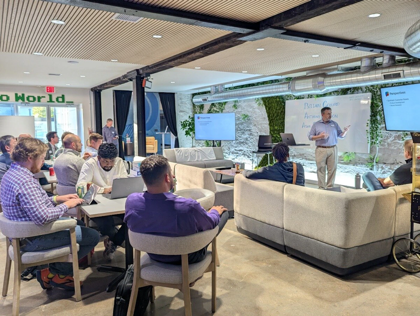 (Photo) A photograph capturing an indoor business meeting or presentation where a speaker stands before a whiteboard and two screens, addressing an audience seated in modern, comfortable lounge furniture. The room features unique architectural elements, including wooden ceiling slats, exposed beams, and a large vertical moss or plant wall. Text: EXIT o World. Perspectives Synthesize Customer [unreadable] Product Development Better Support BRIAN CELLARD AUTHORIZATION A[unreadable] A corporate meeting or training session | Indoor, modern office or event space | A presenter addressing an audience; vertical garden wall; exposed industrial ceiling elements; multiple display screens. | Candid, documentary | Colors: #A9A8A2, #704214, #3E603F, #FFFFFF Note: This image is a realistic capture of a real-world scene (a business meeting), utilizing lighting and perspective characteristic of a photograph rather than a drawn or digitally rendered illustration.