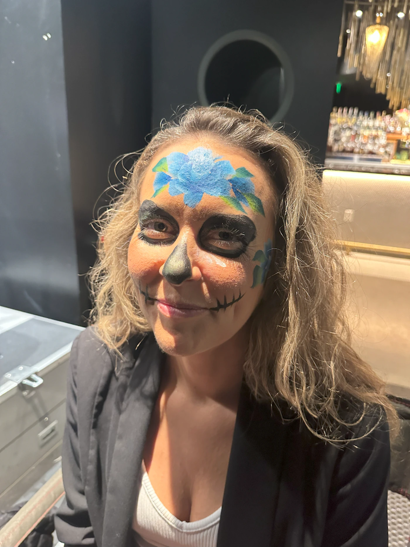 (Photo) A woman with face paint resembling a sugar skull design smiles at the camera. person | indoor | face paint | candid Note: This is a photograph of a person with face paint.  It captures a real-life moment.