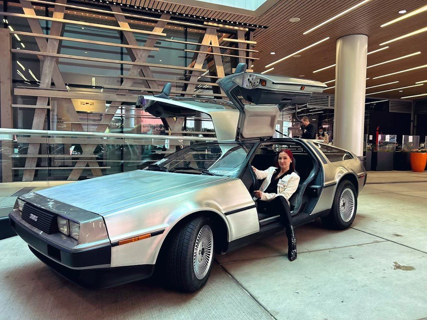 (Photo) A woman is sitting in a DeLorean DMC-12 car with the gull-wing doors open, seemingly posing for a picture. The car is parked indoors in a modern-looking building. car, person | indoor | DeLorean DMC-12 car, gull-wing doors open, person posing | candid Note: This image is a real-life photograph of a car and a person, captured in a specific setting. It's not a drawing, logo, or banner.