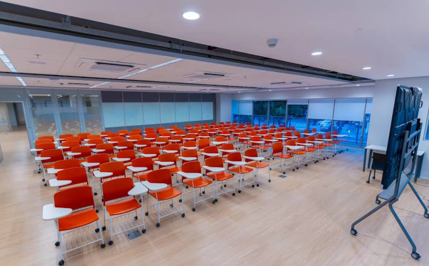 (Photo) A well-lit, modern classroom or lecture hall is shown, featuring numerous rows of vibrant orange chairs with attached white writing tablets. The room has light wooden flooring, large windows with closed blinds on one side, and a large display screen on a mobile stand on the other. Modern classroom or lecture hall | Indoor, educational or corporate facility | Multiple rows of empty orange chairs, light wooden floor, large windows with blinds, a large mobile display screen, bright overhead lighting. | Architectural / Interior photography | Colors: #FF6600, #D4B89A, #E0E0E0, #333333 Note: The image is a realistic capture of an indoor environment, specifically a modern classroom or lecture hall, which is characteristic of a photograph used to document or showcase a physical space.