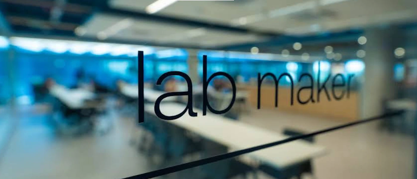 (Photo) A photograph showing the interior of a modern office or laboratory space through a glass partition, with the words 'lab maker' clearly visible in black text on the glass surface. Text: lab maker An interior office or laboratory space with signage on glass. | Indoor, likely a modern workspace or research facility. | Black text 'lab maker' on a transparent glass surface, blurred background showing people and tables, blue lighting or accents in the distant background. | Environmental shot, candid, with shallow depth of field. | Colors: #000000, #005f99, #D3D3D3 Note: The image is a photographic capture of an interior environment, showing a room through a glass partition. The text 'lab maker' is part of the physical environment, appearing as signage on the glass, making it a scene rather than a standalone graphic element.