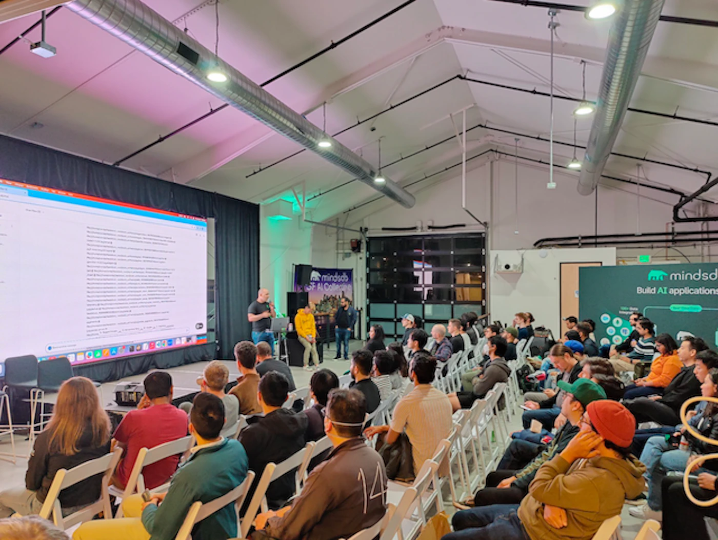 (Photo) A large group of people are seated in an auditorium, watching a presentation displayed on a large screen. Two presenters stand on a stage in front of the screen. Text: mindsdb Build AI applications people | indoor | presentation, audience, presenters | candid Note: The image is a real-life captured scene of an event, depicting people and a presentation, which makes it a photograph.