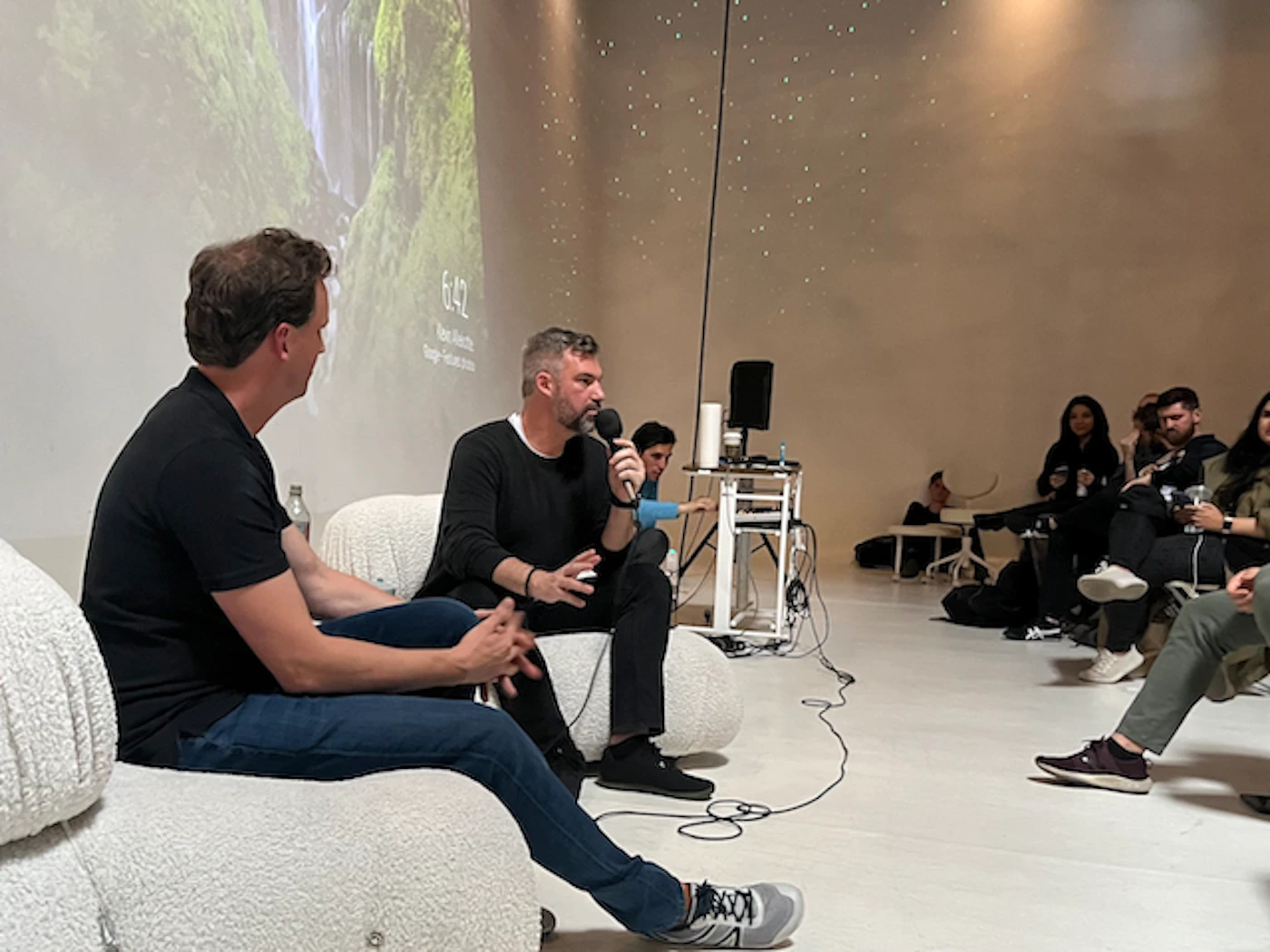 (Photo) Two men are seated on a stage, one holding a microphone, apparently engaging in a discussion or presentation. An audience is seated on chairs in the background. Text: 6:42 Kevin Bethune Design Director people | indoor | presentation, audience, stage | candid Note: The image is a real-life captured scene of an event with people, indicating it's a photograph. It's not staged for artistic or illustrative purposes.
