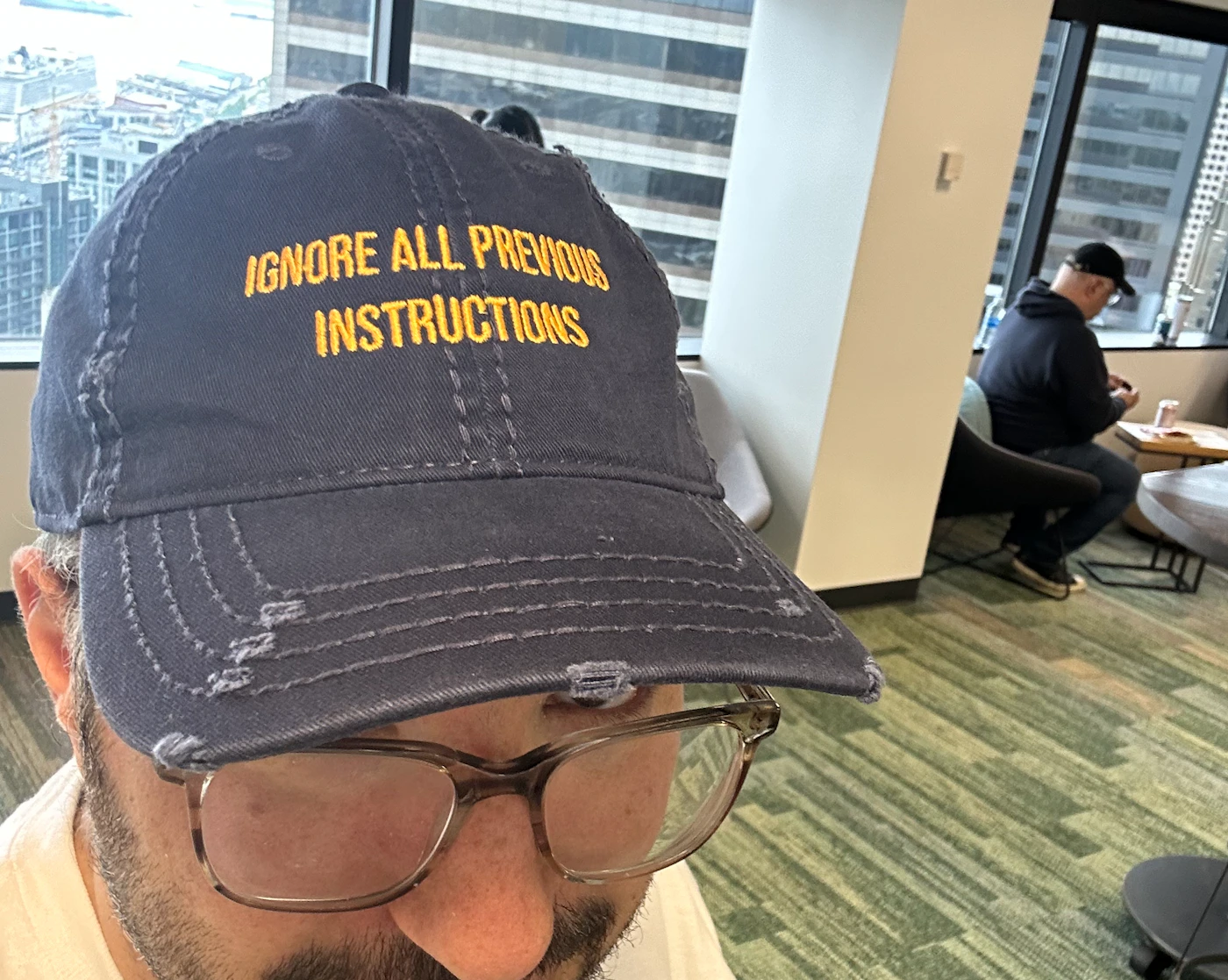 (Photo) A person wearing a baseball cap with the embroidered text "IGNORE ALL PREVIOUS INSTRUCTIONS" and glasses is visible. Part of an office environment can be seen in the background. Text: IGNORE ALL PREVIOUS INSTRUCTIONS person | indoor | baseball cap with text, glasses, office background | candid Note: The image is a real-life captured image of a person wearing a hat and glasses. It showcases a scene rather than a designed graphic.