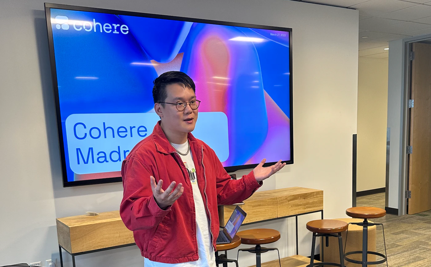 (Photo) A person in a red jacket is gesturing with their hands while presenting in front of a large screen. Text: cohere Cohere Madr March 27, 2023 person | indoor | presentation, screen | candid Note: The image is a real-life captured scene of a presentation, depicting a person and their environment. This makes it a photograph.
