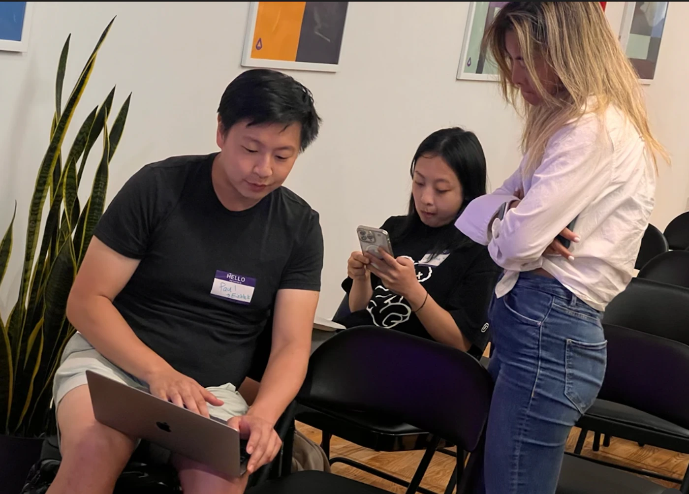(Photo) Three people are in a room with folding chairs. One person is using a laptop, another is on their phone, and the third person is standing with their arms crossed. Text: HELLO Paul [unreadable] people | indoor | multiple people, laptop, smartphone | candid Note: The image is a candid photograph of people in a room. It captures a real-life scene rather than a designed graphic or illustration.