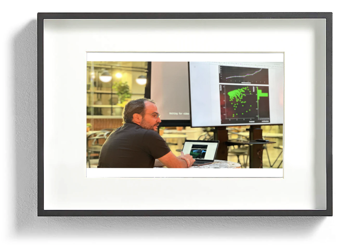 (Photo) A framed photograph of a man sitting at a table with a laptop, looking at a presentation displayed on screens. person | indoor | laptop, presentation screens | candid Note: The image is a real-life captured image of a person in an indoor setting, indicative of a photograph.  It's not a drawing, logo, or other graphical representation.