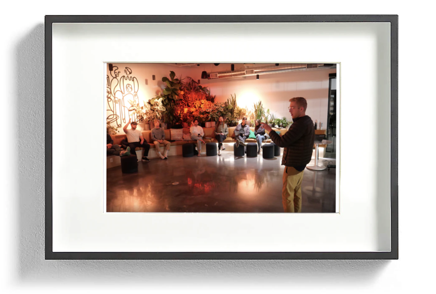 (Photo) A framed photograph of a person presenting or leading a discussion with a group of people seated in a room. people | indoor | group of people, presentation/meeting | candid Note: The image is a realistic depiction of a scene captured by a camera, showing a group of people in a room with one person seemingly leading a discussion.  It is a framed photograph of this scene.