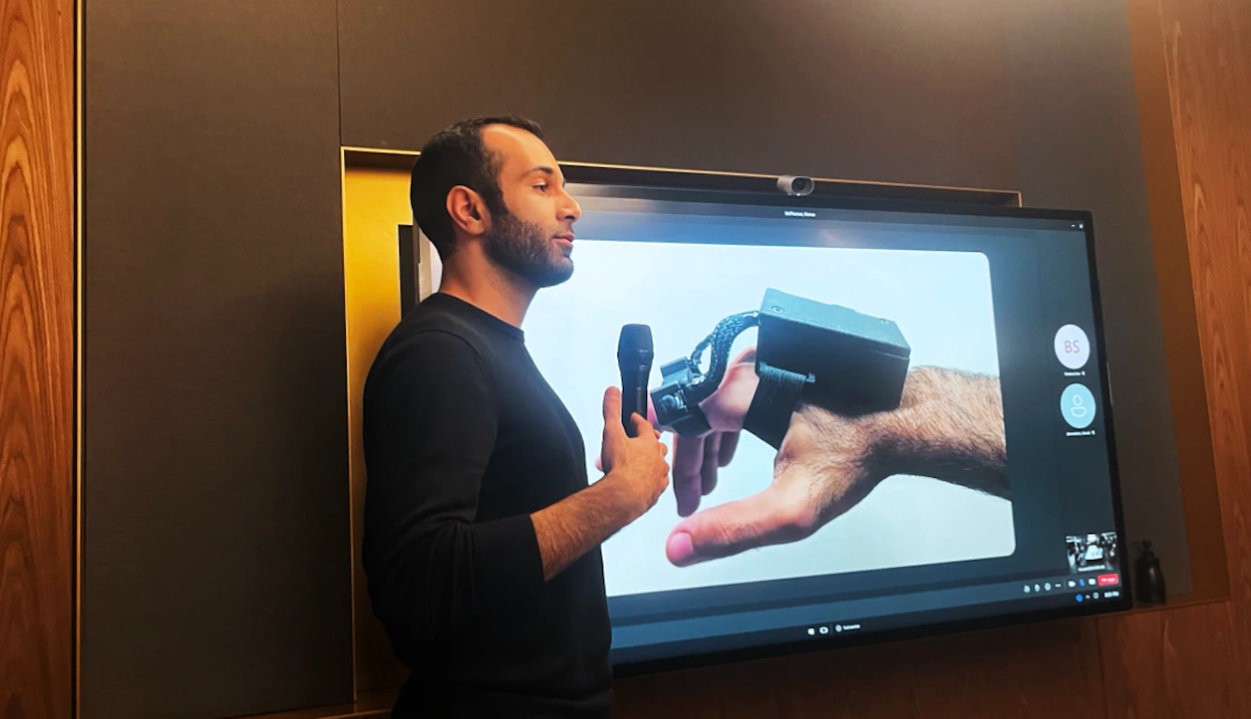 (Photo) A man stands in profile holding a microphone, presenting to a screen displaying an image of a hand with a device attached. Text: 85 person | indoor | presentation, screen, technology | candid Note: The image is a real-life captured moment of a presentation, making it a photograph. It depicts a person and a screen, not a logo, illustration, or other specific type.